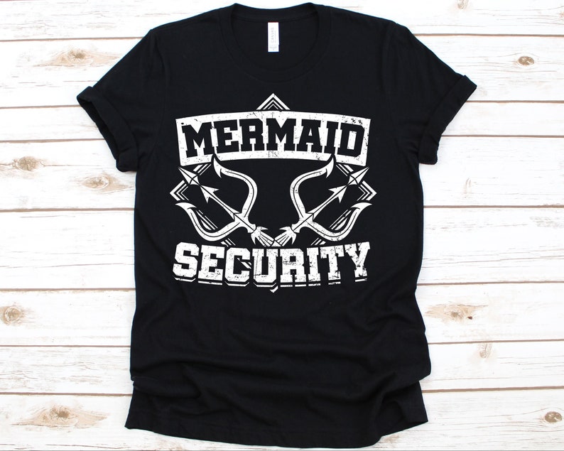Puede incluir: Camiseta negra con la frase "MERMAID IS SECURITY" en blanco con tipograf&iacute;a desgastada. El dise&ntilde;o incluye dos gr&aacute;ficos de tridente. La camiseta es de material suave y tiene cuello redondo.