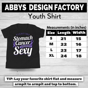 Op de afbeelding: Zwart jeugdshirt met de tekst "FIGHTING Stomach Cancer AND STILL THIS Sexy" in wit en paars. Afmetingen in inches worden gegeven voor de maten S, M, L en XL. Het shirt is van Abbys Design Factory.