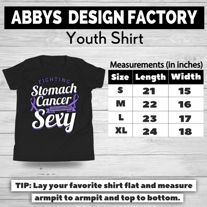 Op de afbeelding: Zwart jeugdshirt met de tekst "FIGHTING Stomach Cancer AND STILL THIS Sexy" in wit en paars. Afmetingen in inches worden gegeven voor de maten S, M, L en XL. Het shirt is van Abbys Design Factory.