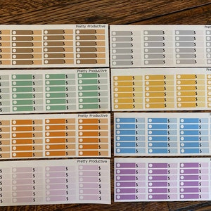 May include: A collection of colorful planner stickers with the text "Pretty Productive." Each sticker features a series of boxes with dollar signs and circles. The stickers are in various colors, including brown, gray, green, orange, blue, and purple.