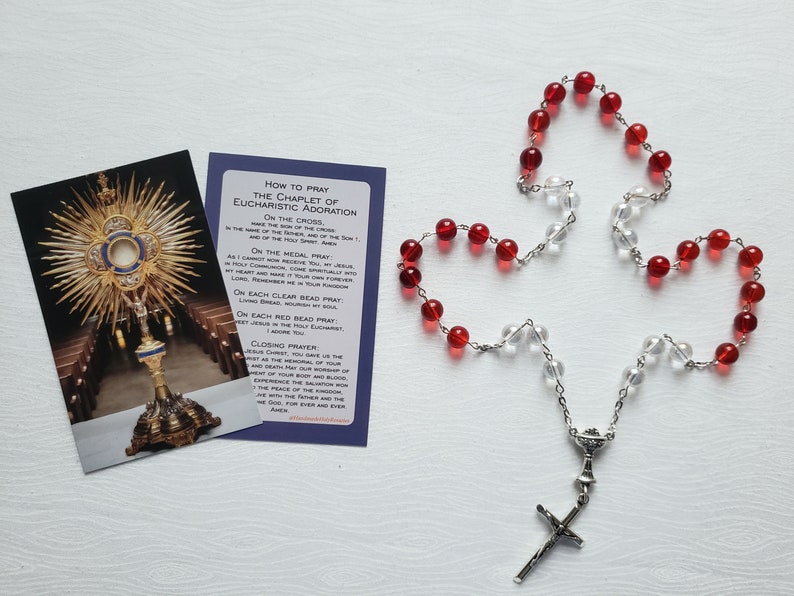 Pu&ograve; includere: Un rosario d'argento con perline di vetro rosso e trasparente e un crocifisso d'argento. &Egrave; anche raffigurata una carta con le istruzioni su come pregare il chaplet di Adorazione Eucaristica.