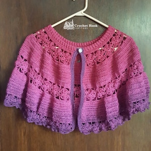 May include: A handmade, open-front crochet shrug in shades of pink and purple, hanging on a white hanger. The shrug features a scalloped edge and decorative floral patterns. The Crochet Hook Cottage logo is visible on the hanger.