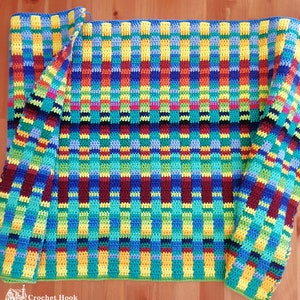 Puede incluir: Una manta de ganchillo hecha a mano con un patrón vibrante y colorido. La manta presenta un diseño tejido con rayas verticales azules, verdes, amarillas, naranjas y rojas. La manta está doblada, revelando el intrincado trabajo de puntadas y la textura.