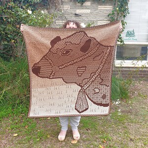May include: A rectangular crochet blanket featuring a large, dark brown profile of a badger head against a light tan background. The design incorporates intricate geometric patterns and a Greek key border in dark brown and light tan yarn colors.
