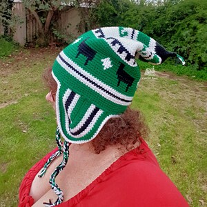 May include: A green, white, and black crochet hat with ear flaps and a long, pointed top. The hat features a pattern of black sheep and white geometric shapes. The hat is handmade and has braided ties with green, white, and black strands.