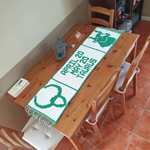 Könnte beinhalten: Weiß-grüner gehäkelter Tischläufer mit dem Text "APRIL 20 MAY 20 TAURUS" und einem Stier-Symbol. Der Läufer liegt auf einem Holztisch, zusammen mit einem Buch und einer kleinen Statue.