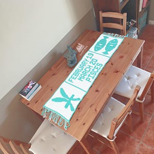May include: A white and turquoise crochet table runner with the text "FEBRUARY 19 MARCH 20 PISCES" and Pisces symbols. The runner is fringed and sits on a wooden table. A small gray elephant statue and a book are also on the table.