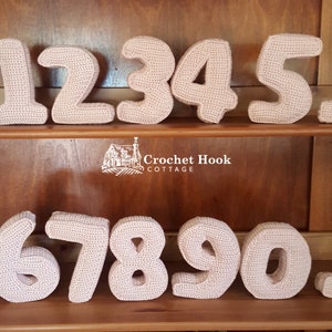 May include: Crocheted numbers 1 through 10 in a light pink color. The numbers are arranged in two rows on a wooden shelf. The shelf has a sign that reads "Crochet Hook Cottage".