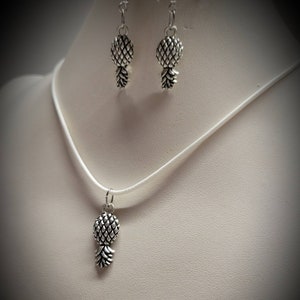 May include: A silver pineapple necklace and matching earrings. The necklace has a white cord and the earrings have silver hooks.