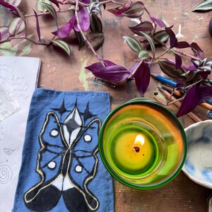 May include: A lit candle in a green glass holder sits next to a blue fabric item with a butterfly design. The butterfly is black, white, and gold. Purple and green leaves and stems are in the background, along with colored pencils and a small bowl.
