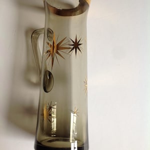 Puede incluir: Una jarra alta de cristal gris ahumado con un borde dorado y un patrón repetitivo de estrellas doradas. La jarra tiene un asa de cristal transparente en el lateral y una silueta cónica con un pico vertedor ancho y curvado en la parte superior.