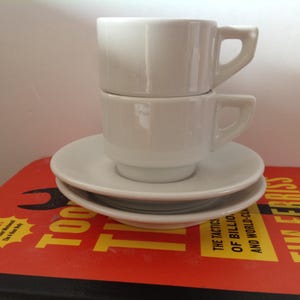 May include: Two white ceramic coffee cups stacked on top of each other, each with a handle. They sit on a stack of two matching white saucers. The cups and saucers are on a red and yellow book.