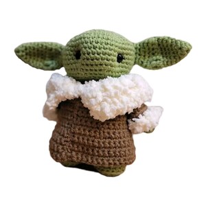 May include: A handmade crochet doll in the likeness of a popular character. The doll features large, pointed ears, a green head, and a brown robe with a white fluffy collar. The doll has black eyes and is made of yarn.