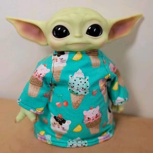 May include: A green-skinned doll with large black eyes and large ears, wearing a turquoise shirt. The shirt is patterned with ice cream cones, cat faces, cherries, and lemon slices. The doll is a popular character from a science fiction franchise.