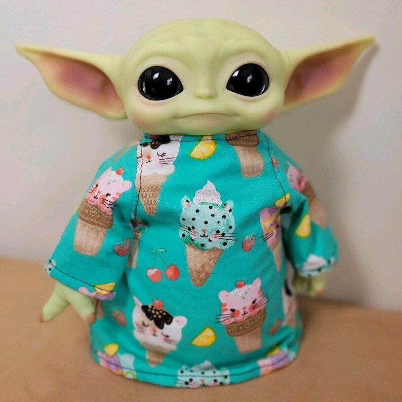 May include: A green-skinned doll with large black eyes and large ears, wearing a turquoise shirt. The shirt is patterned with ice cream cones, cat faces, cherries, and lemon slices. The doll is a popular character from a science fiction franchise.