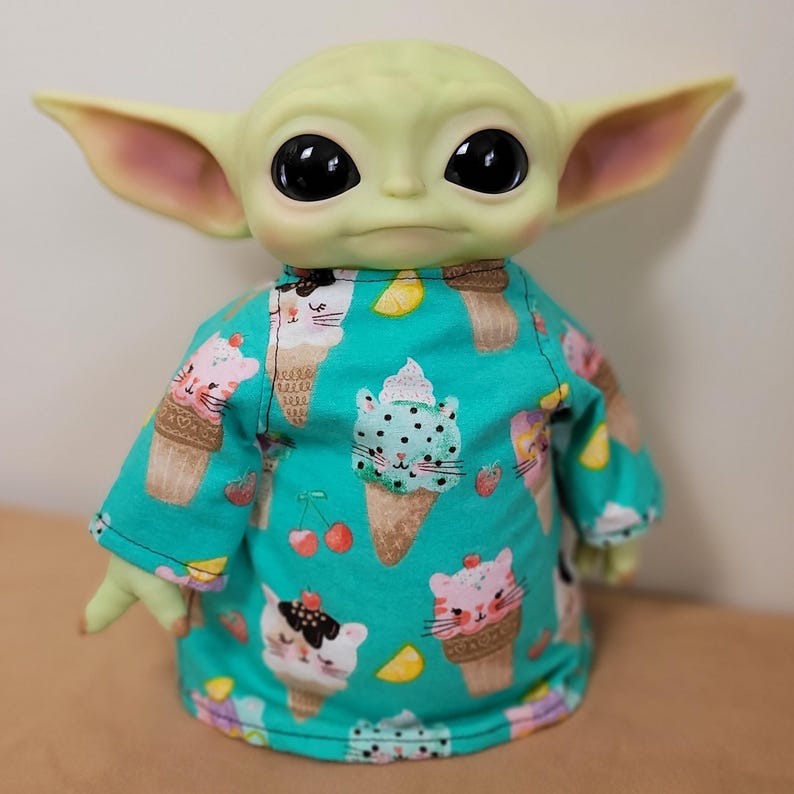 May include: A doll of a character with large black eyes and pointed ears, wearing a turquoise shirt. The shirt is patterned with ice cream cones, cats, cherries, and lemon slices. The doll has a light green complexion.