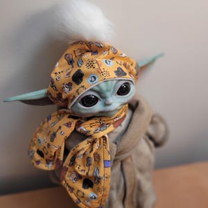 Puede incluir: Mu&ntilde;eco de Grogu, tambi&eacute;n conocido como Baby Yoda, con un gorro amarillo con un pomp&oacute;n blanco y una bufanda a juego. El gorro y la bufanda tienen un estampado de instrumentos musicales. El mu&ntilde;eco tiene grandes ojos negros y piel verde.