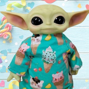 May include: A doll of a character with large ears and black eyes, wearing a teal onesie. The onesie is patterned with ice cream cones and cat faces. The background includes a lollipop, confetti, and a light blue wooden surface.