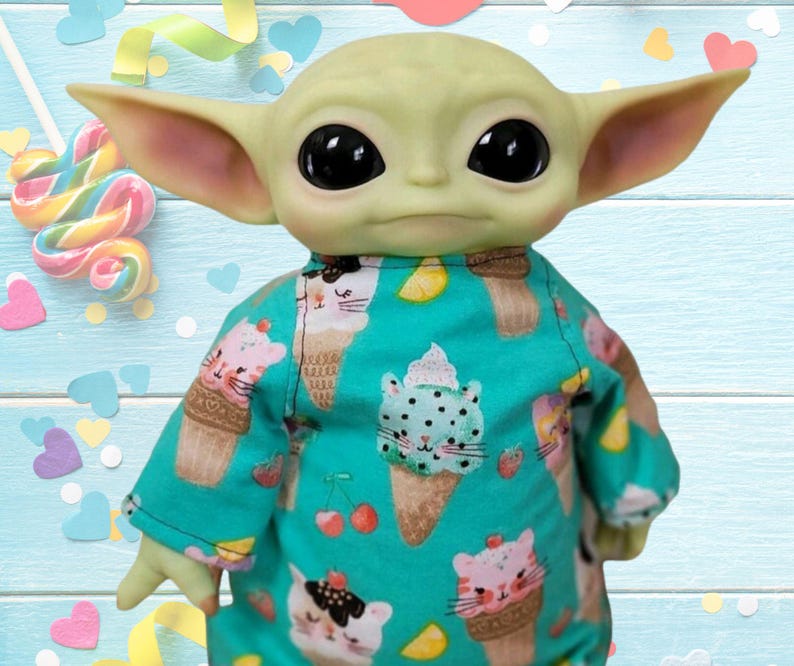 May include: A doll of a character with large ears and black eyes, wearing a teal onesie. The onesie is patterned with ice cream cones and cat faces. The background includes a lollipop, confetti, and a light blue wooden surface.