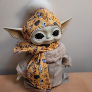 Puede incluir: Mu&ntilde;eco de un personaje de una popular serie de ciencia ficci&oacute;n, con un gorro y bufanda amarillos estampados. El gorro tiene un pomp&oacute;n blanco. El mu&ntilde;eco lleva un abrigo gris con cuello de piel beige.