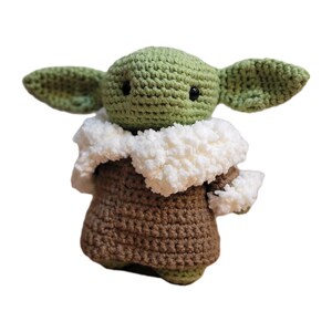 May include: A crocheted figure of a character with large ears, green skin, and a brown robe. The figure has a white, fluffy collar and black eyes. The handmade item is a soft sculpture.