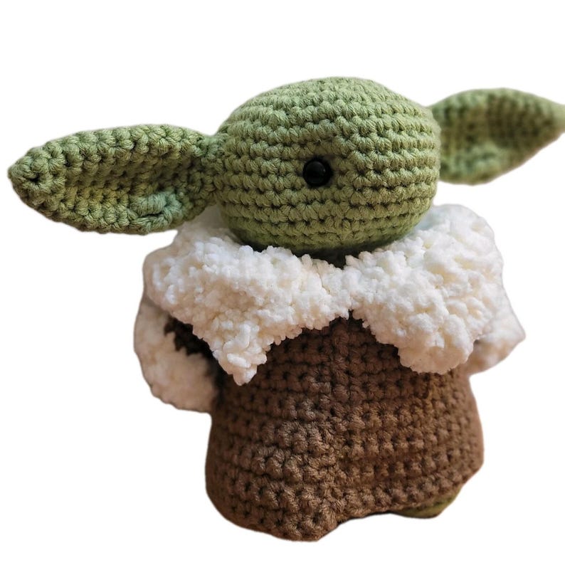 May include: A handmade crochet doll of a character with large ears and a green head. The doll has a brown body, a white fluffy collar, and black eyes. The doll is a popular character from a science fiction franchise.