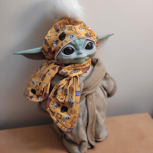 Puede incluir: Mu&ntilde;eco Grogu con un gorro y bufanda amarillos con un estampado de instrumentos musicales. El gorro tiene un pomp&oacute;n blanco. El mu&ntilde;eco lleva una bata color beige.