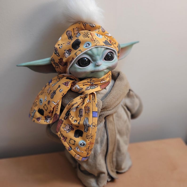 Puede incluir: Mu&ntilde;eco Grogu con un gorro y bufanda amarillos con un estampado de instrumentos musicales. El gorro tiene un pomp&oacute;n blanco. El mu&ntilde;eco lleva una bata color beige.