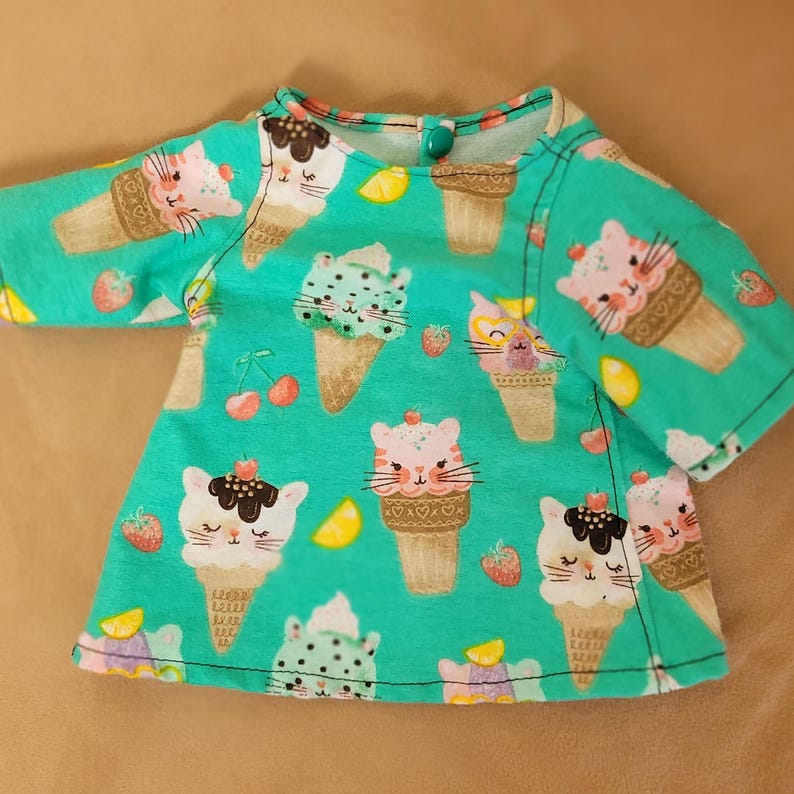 May include: A turquoise-colored doll's shirt with a pattern of ice cream cones with cat faces, strawberries, cherries, and lemon slices. The shirt has a round neckline and short sleeves. A single green button is at the back of the neck.