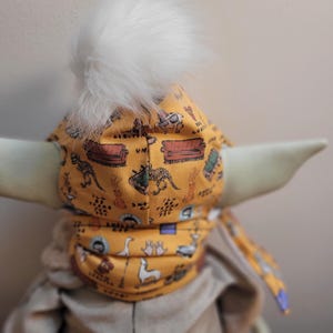 Puede incluir: Un gorro de beb&eacute; amarillo mostaza con un pomp&oacute;n blanco y esponjoso. El gorro est&aacute; estampado con im&aacute;genes de dibujos animados de un sof&aacute;, dinosaurios y otros objetos. El gorro est&aacute; atado con una tela a juego.