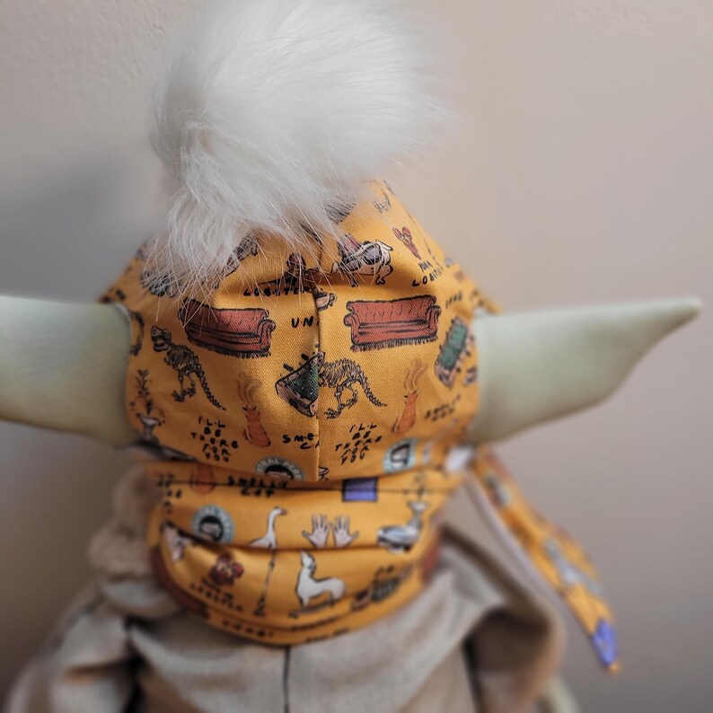 Puede incluir: Un gorro de beb&eacute; amarillo mostaza con un pomp&oacute;n blanco y esponjoso. El gorro est&aacute; estampado con im&aacute;genes de dibujos animados de un sof&aacute;, dinosaurios y otros objetos. El gorro est&aacute; atado con una tela a juego.