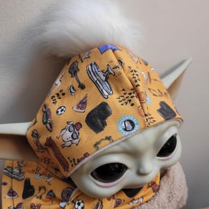 Puede incluir: Una mu&ntilde;eca Grogu con un gorro y bufanda de color amarillo mostaza. El gorro tiene un pomp&oacute;n blanco y est&aacute; estampado con varias ilustraciones, incluyendo una guitarra, una porci&oacute;n de pizza y un perro. La bufanda tambi&eacute;n est&aacute; estampada con las mismas ilustraciones.