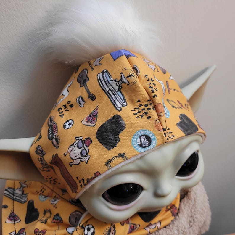 Puede incluir: Una mu&ntilde;eca Grogu con un gorro y bufanda de color amarillo mostaza. El gorro tiene un pomp&oacute;n blanco y est&aacute; estampado con varias ilustraciones, incluyendo una guitarra, una porci&oacute;n de pizza y un perro. La bufanda tambi&eacute;n est&aacute; estampada con las mismas ilustraciones.