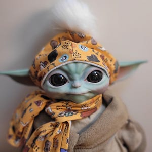 Puede incluir: Mu&ntilde;eco de un personaje de una popular franquicia de ciencia ficci&oacute;n, que lleva un gorro y una bufanda amarillos con un patr&oacute;n repetido de instrumentos musicales y otras formas. El mu&ntilde;eco tiene grandes ojos negros, piel verde y un pomp&oacute;n blanco y esponjoso en el gorro.