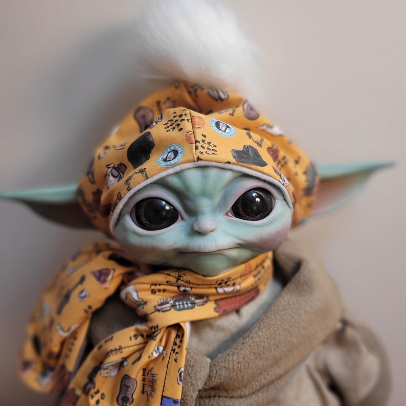 Puede incluir: Mu&ntilde;eco de un personaje de una popular franquicia de ciencia ficci&oacute;n, que lleva un gorro y una bufanda amarillos con un patr&oacute;n repetido de instrumentos musicales y otras formas. El mu&ntilde;eco tiene grandes ojos negros, piel verde y un pomp&oacute;n blanco y esponjoso en el gorro.