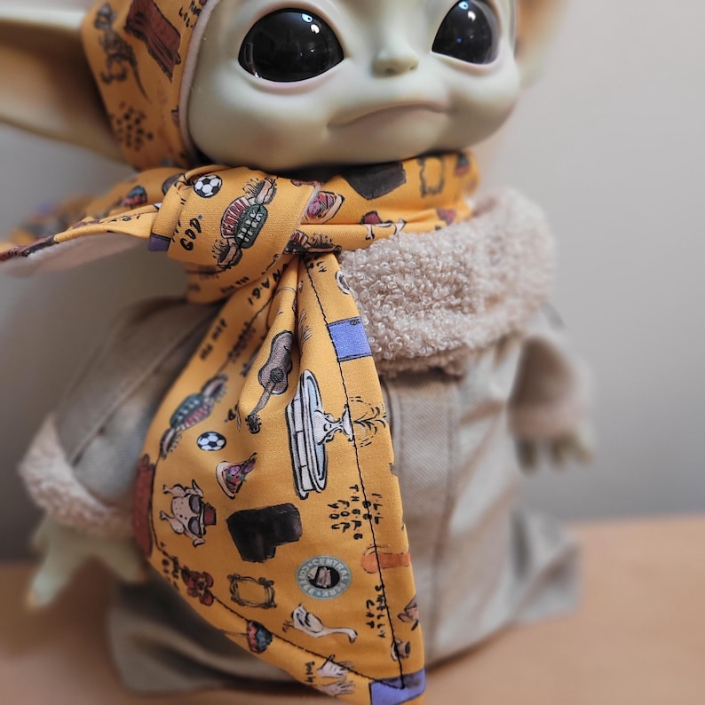 Puede incluir: Mu&ntilde;eco de un personaje de una popular serie de ciencia ficci&oacute;n, con una bufanda amarilla y un gorro a juego. La bufanda tiene un estampado con varias ilustraciones y texto. El mu&ntilde;eco lleva un abrigo beige con un cuello peludo.
