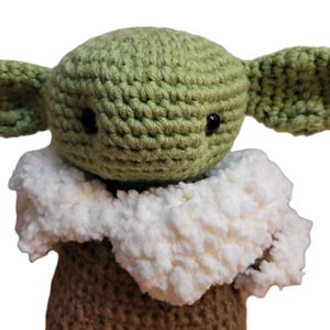 May include: A handmade crochet doll featuring a green head with large ears and black eyes. The doll has a white, fluffy collar and a brown body. The doll is a character from a popular science fiction franchise.