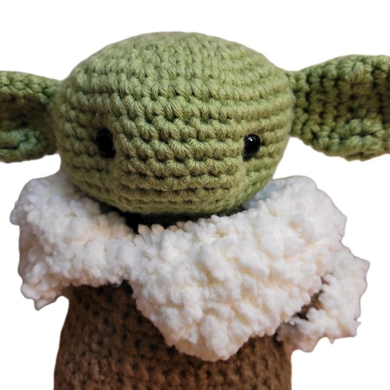 May include: A handmade crochet doll featuring a green head with large ears and black eyes. The doll has a white, fluffy collar and a brown body. The doll is a character from a popular science fiction franchise.
