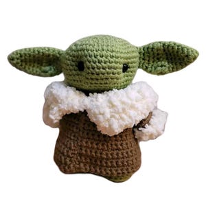 May include: A crocheted doll of a character with large ears, a green head, and a brown body. The doll has black eyes and a white fluffy collar. The doll is handmade and appears to be a character from a popular science fiction franchise.