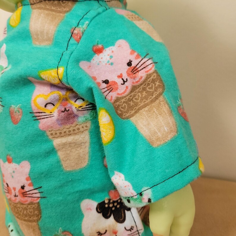 May include: A turquoise fabric with a repeating pattern of ice cream cone cats, strawberries, and lemon slices. The cats have pink fur and wear sunglasses. The fabric is sewn into a garment.