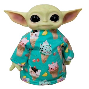 May include: A green-skinned doll with large black eyes and pointed ears, wearing a turquoise shirt. The shirt is patterned with ice cream cones and cat faces. The doll has small hands and is set against a white background.