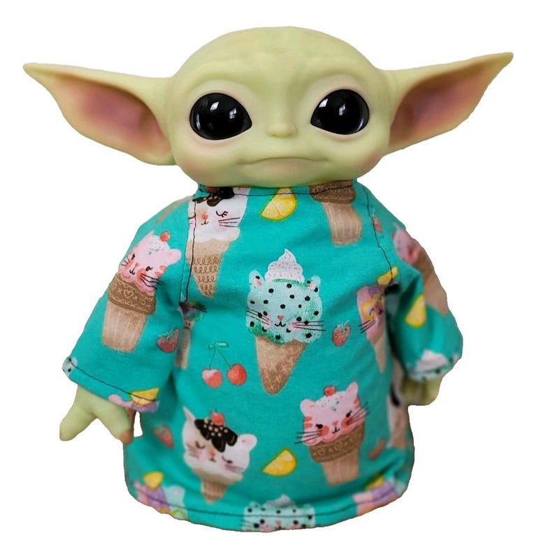 May include: A green-skinned doll with large black eyes and pointed ears, wearing a turquoise shirt. The shirt is patterned with ice cream cones and cat faces. The doll has small hands and is set against a white background.