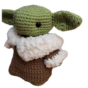 May include: Crocheted figure of a character with large ears, green skin, and black eyes. The figure is wearing a brown and white outfit. The handmade item is a plush toy, likely inspired by a popular science fiction character.