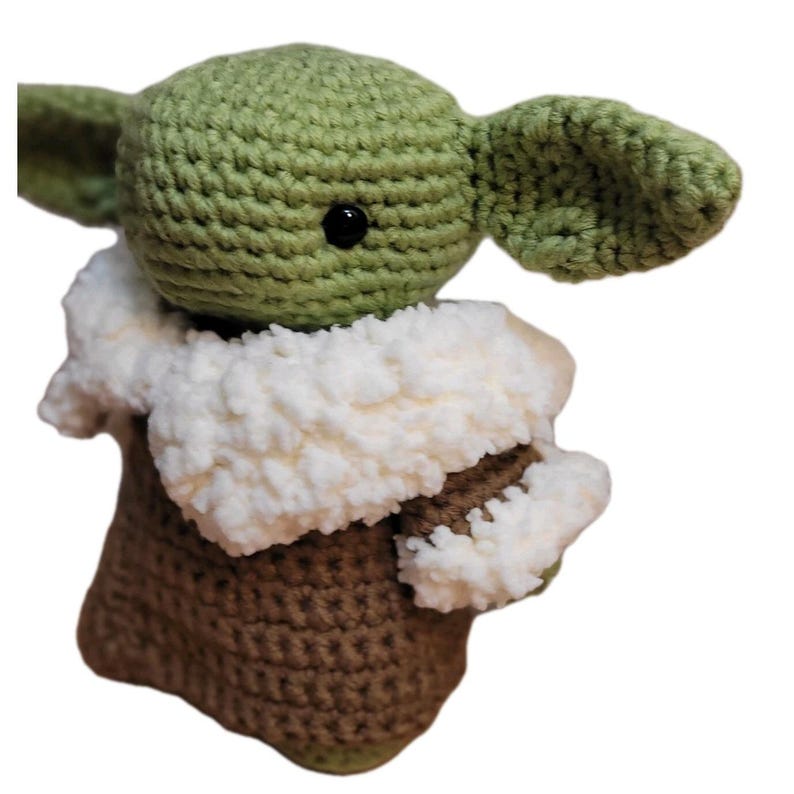 May include: Crocheted figure of a character with large ears, green skin, and black eyes. The figure is wearing a brown and white outfit. The handmade item is a plush toy, likely inspired by a popular science fiction character.