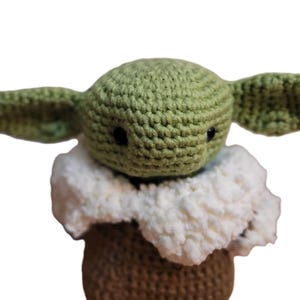 May include: Crocheted doll of a character with large ears, a green head, and black eyes. The doll has a white, fluffy collar and a brown base. The handmade toy is a popular character from a science fiction series.