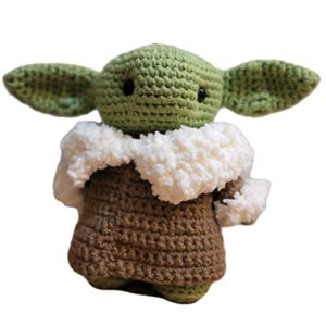 May include: A handmade crochet doll of a character with large green ears, black eyes, and a brown robe. The doll has a white fluffy collar and is made of green and brown yarn. The doll is standing upright against a white background.