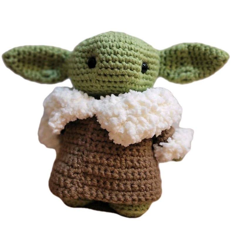 May include: A handmade crochet doll of a character with large green ears, black eyes, and a brown robe. The doll has a white fluffy collar and is made of green and brown yarn. The doll is standing upright against a white background.