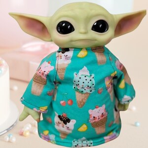 May include: A figurine of a character with large ears and black eyes, wearing a teal shirt. The shirt is patterned with ice cream cones with cat faces, cherries, and strawberries. The figurine is light green and has a neutral expression.
