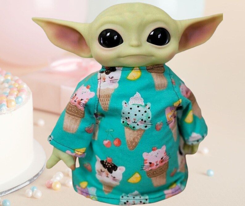 May include: A figurine of a character with large ears and black eyes, wearing a teal shirt. The shirt is patterned with ice cream cones with cat faces, cherries, and strawberries. The figurine is light green and has a neutral expression.
