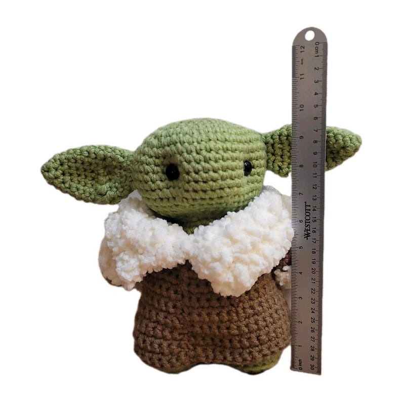 May include: A handmade crochet doll of a character with large ears, black eyes, and a brown robe. The doll is approximately 6 inches tall, with a white fluffy collar. A ruler is placed next to the doll for size comparison.